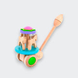 Wooden carousel push toy with horse figures, colorful wheels, and a long handle, shown at an angle on a light gray background.