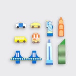 New York City-themed wooden toy pieces laid out separately on a light gray background: a police car, yellow taxi, school bus, hot dog cart, blue bridge, Statue of Liberty, and three colorful buildings.⠋
