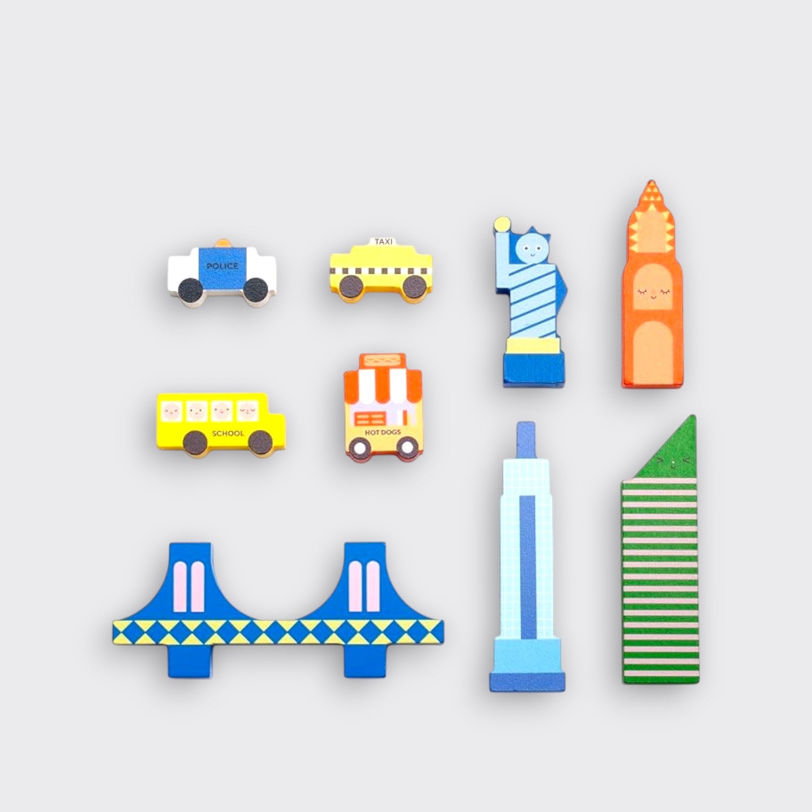 New York City-themed wooden toy pieces laid out separately on a light gray background: a police car, yellow taxi, school bus, hot dog cart, blue bridge, Statue of Liberty, and three colorful buildings.⠋