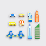 New York City-themed wooden toy pieces laid out separately on a light gray background: a police car, yellow taxi, school bus, hot dog cart, blue bridge, Statue of Liberty, and three colorful buildings.⠋