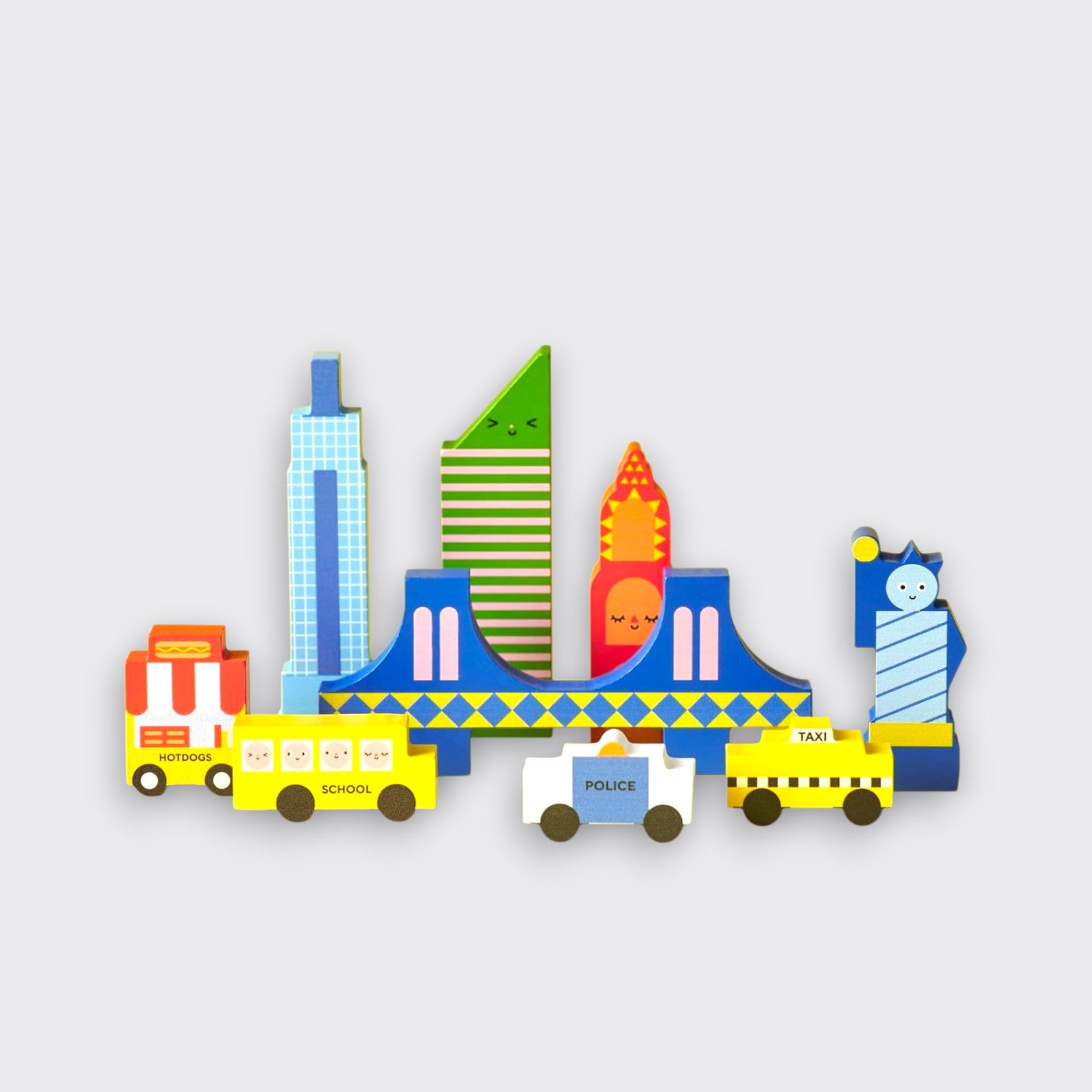 Colorful New York City-themed wooden toy pieces arranged on a light gray background. Skyscrapers and a blue bridge stand behind a hot dog cart, school bus, police car, yellow taxi, and a stylized Statue of Liberty.